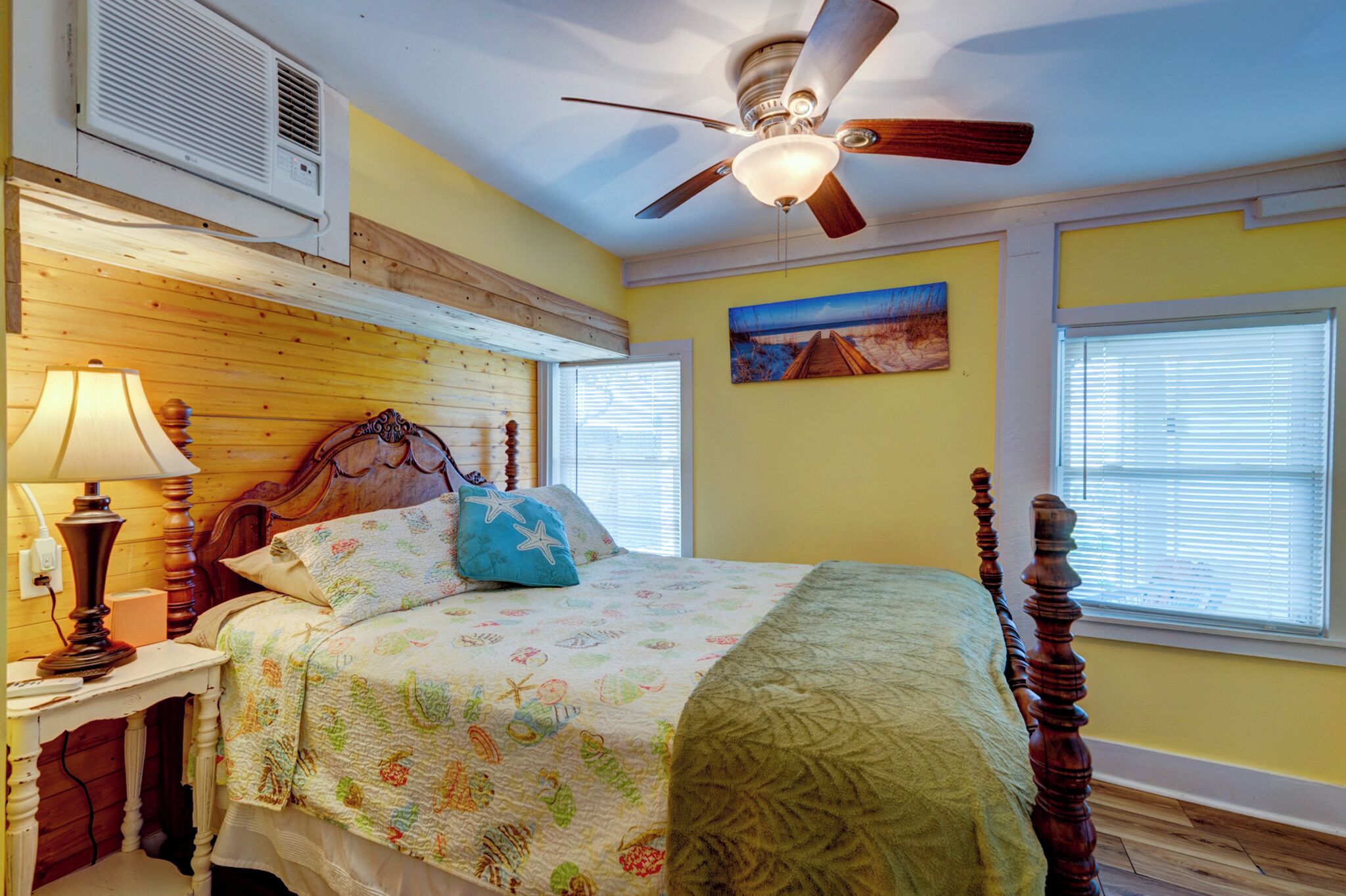 A bedroom with a wooden headboard, a ceiling fan, a bed with floral bedding, a nightstand with a lamp, and a painting on the wall.