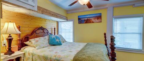 A bedroom with a wooden headboard, a ceiling fan, a bed with floral bedding, a nightstand with a lamp, and a painting on the wall.