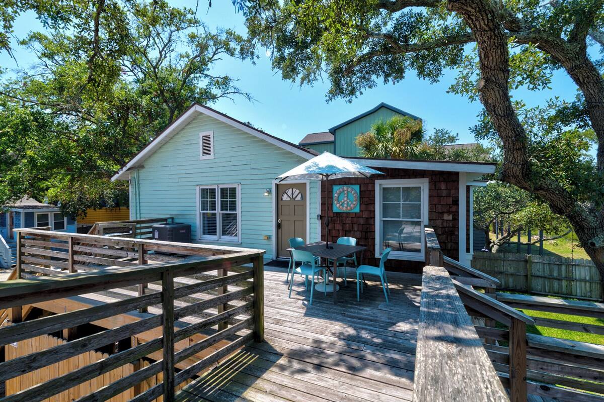 A wooden deck with a table and chairs, a small brick structure with a clock, and a light blue house in the background.