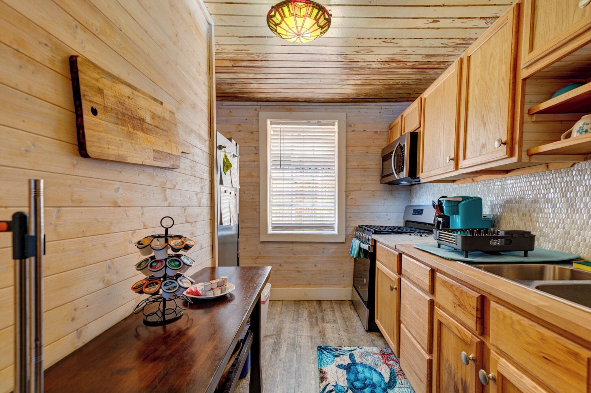 A compact kitchen with wooden walls, a wooden ceiling, a window with blinds, a microwave, a stove, and a coffee maker.