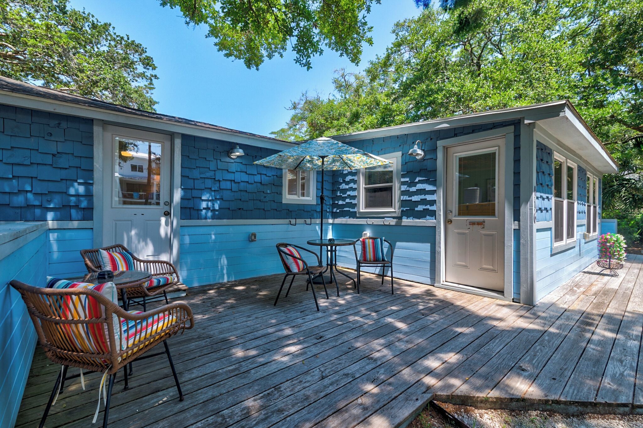 A blue house with a wooden deck, two wicker chairs, a small table, and a blue umbrella.