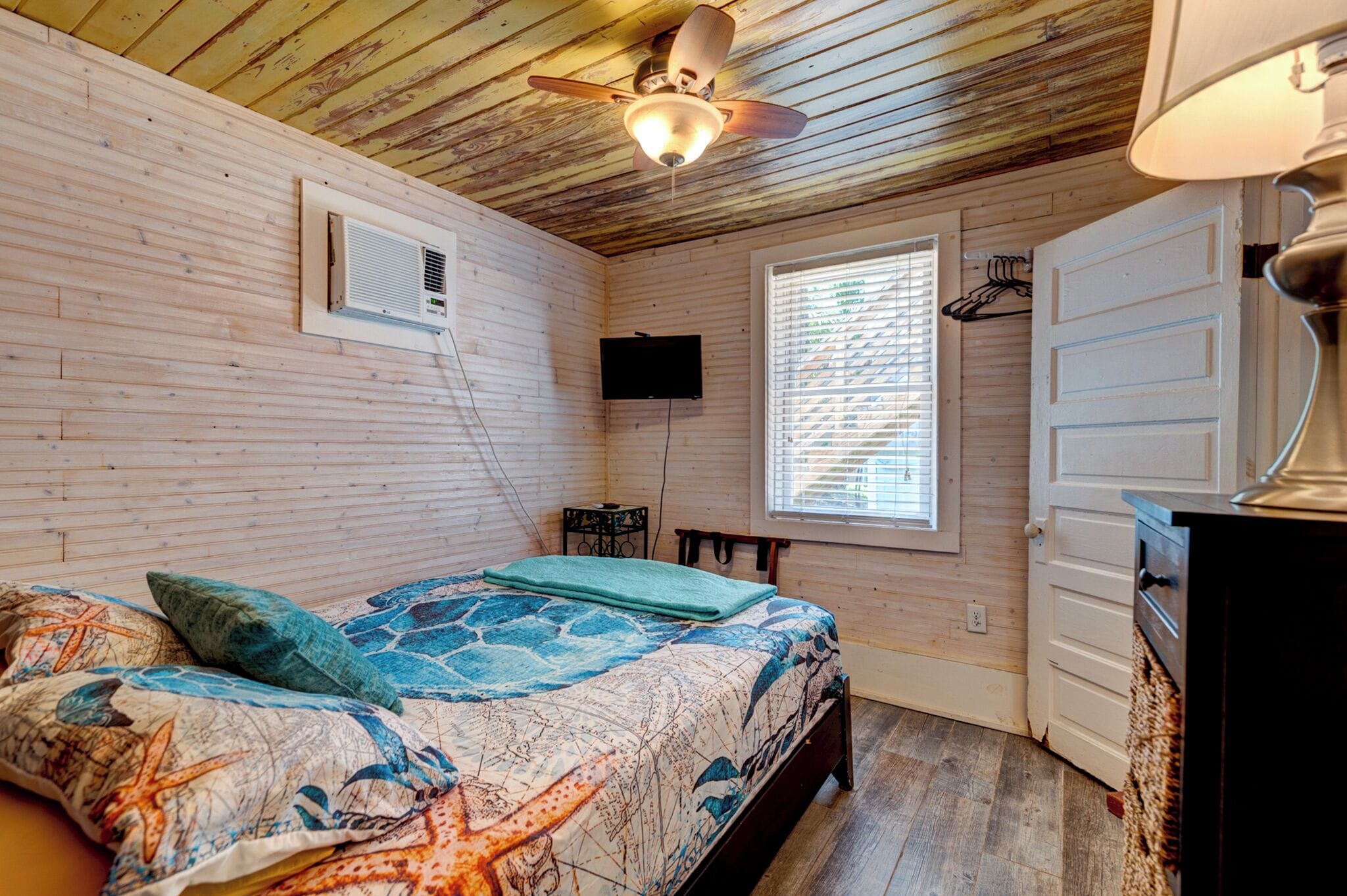 Een slaapkamer met een houten plafond, een bed met een beddegoed met een patroon, een plafondventilator, een aan de muur bevestigde televisie en een raam met jaloezieën.