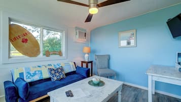 Una sala de estar con un sofá azul, una mesa de centro blanca, un ventilador de techo y un televisor sobre una mesa blanca.
