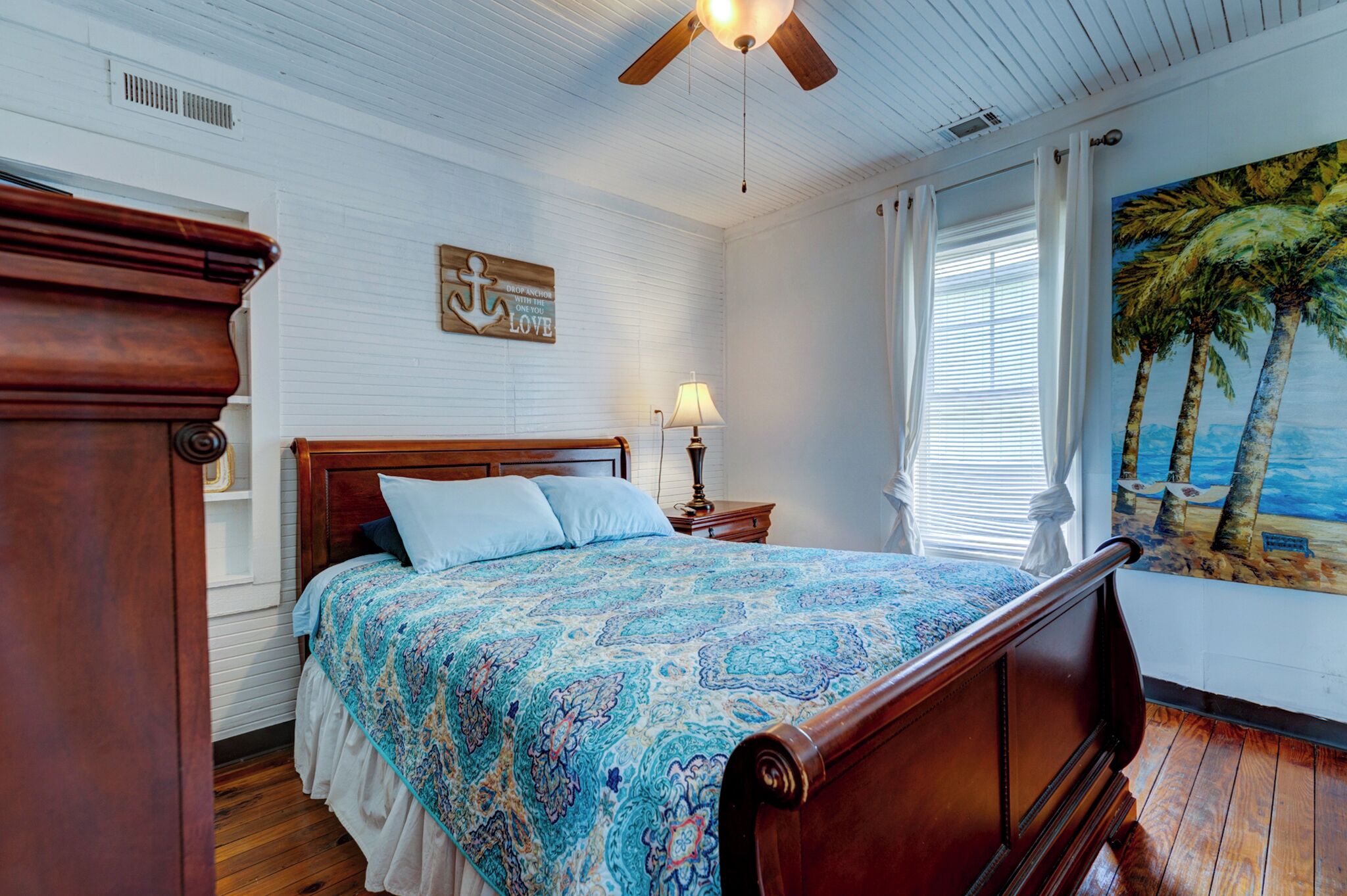 A bedroom with a wooden bed, a nightstand with a lamp, a ceiling fan, and a painting of palm trees.