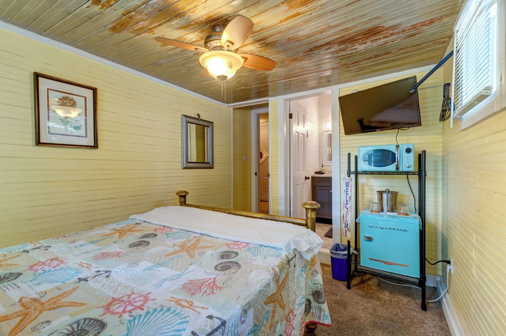 Una habitación con cama, ventilador de techo, televisor y refrigerador antiguo.