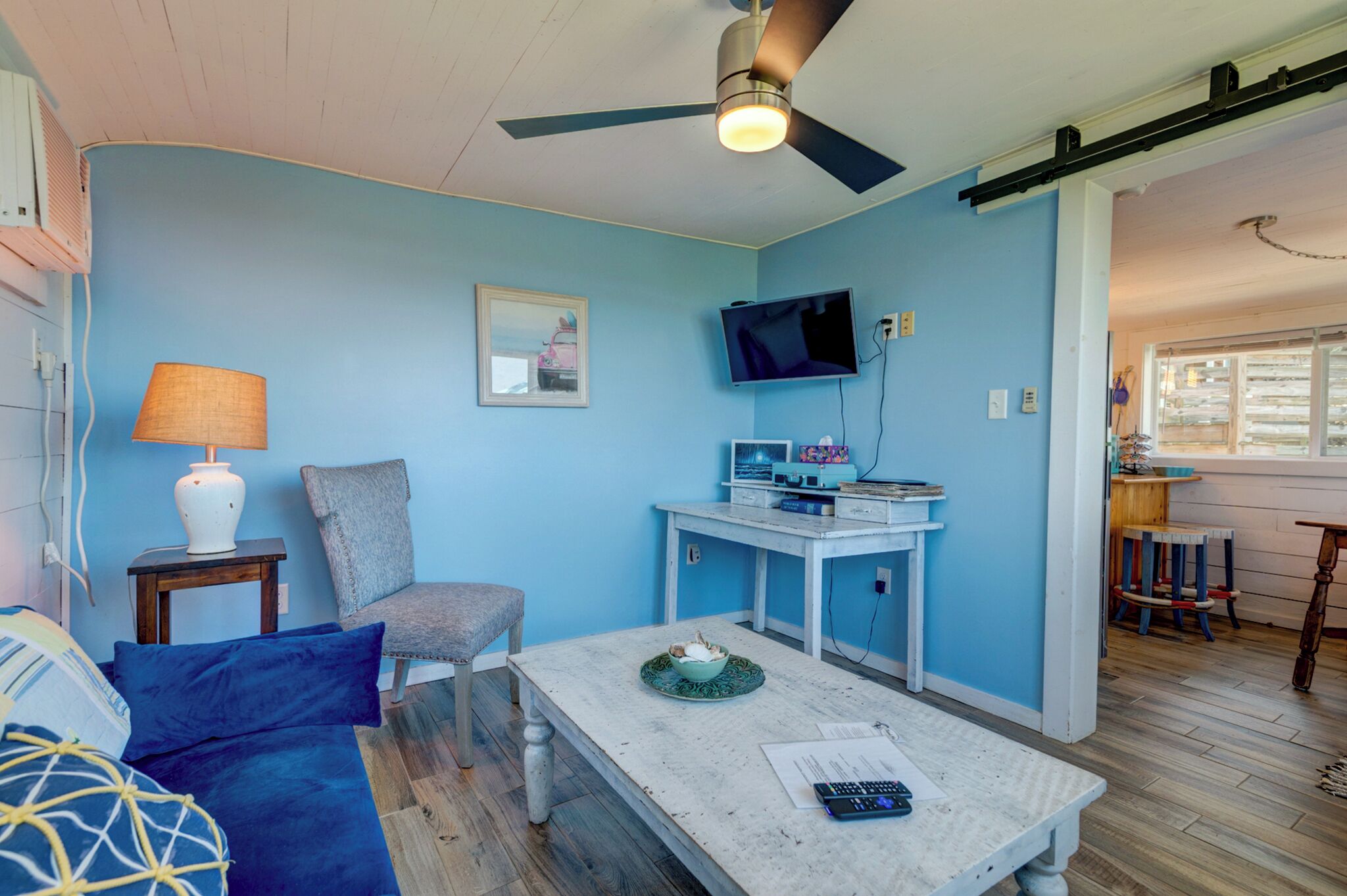 Een kamer met een blauwe muur, een plafondventilator, een aan de muur bevestigde televisie, een kleine tafel met een kom en papieren, een stoel en een bed met kussens.