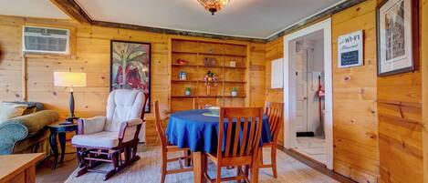 Una habitación acogedora con paredes de madera, una mesa de comedor con sillas, un sillón mecedor, una lámpara y una estantería.