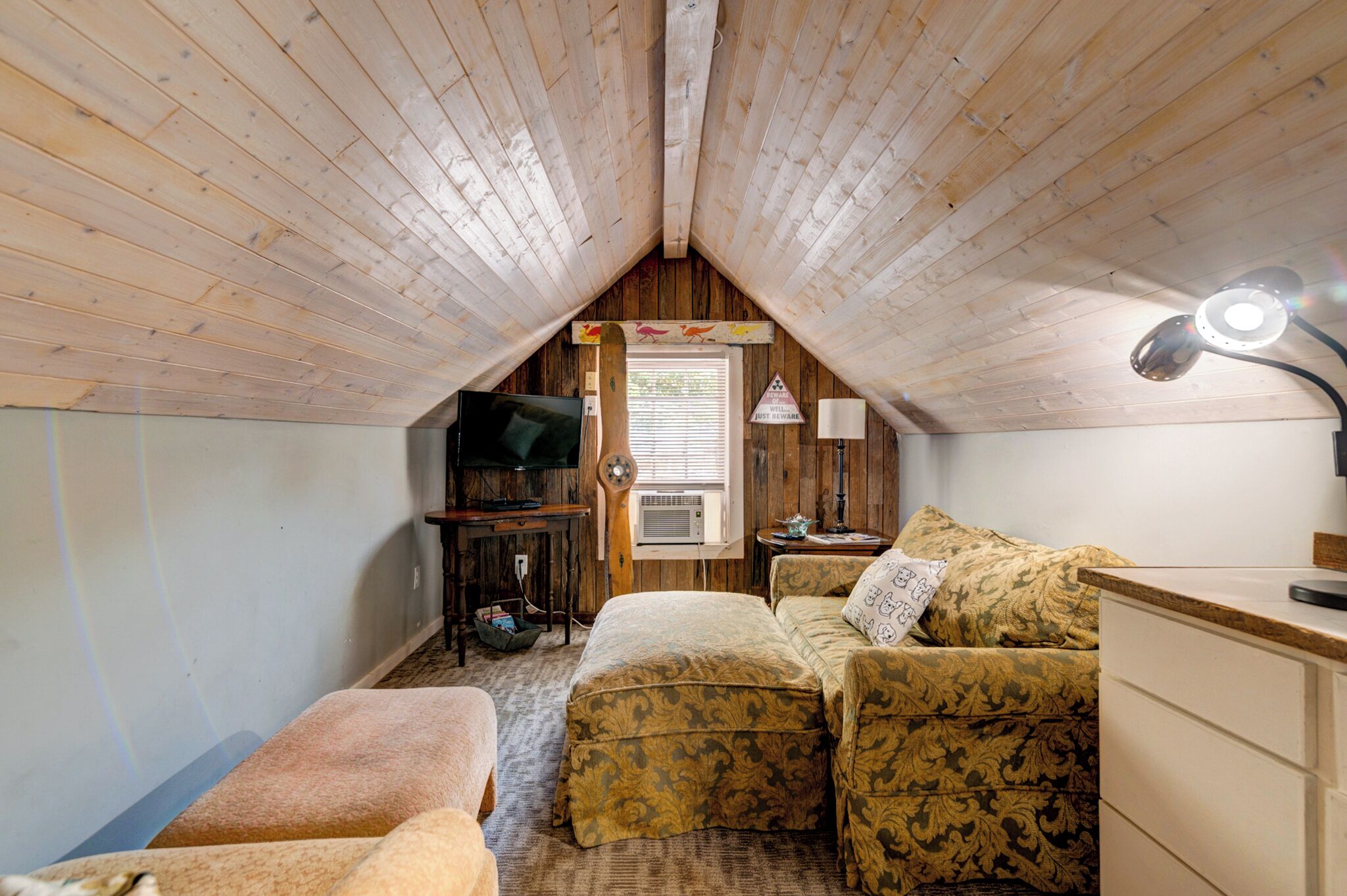 Acogedor dormitorio en ático con techo de madera inclinado, televisión, lámpara y sofá.