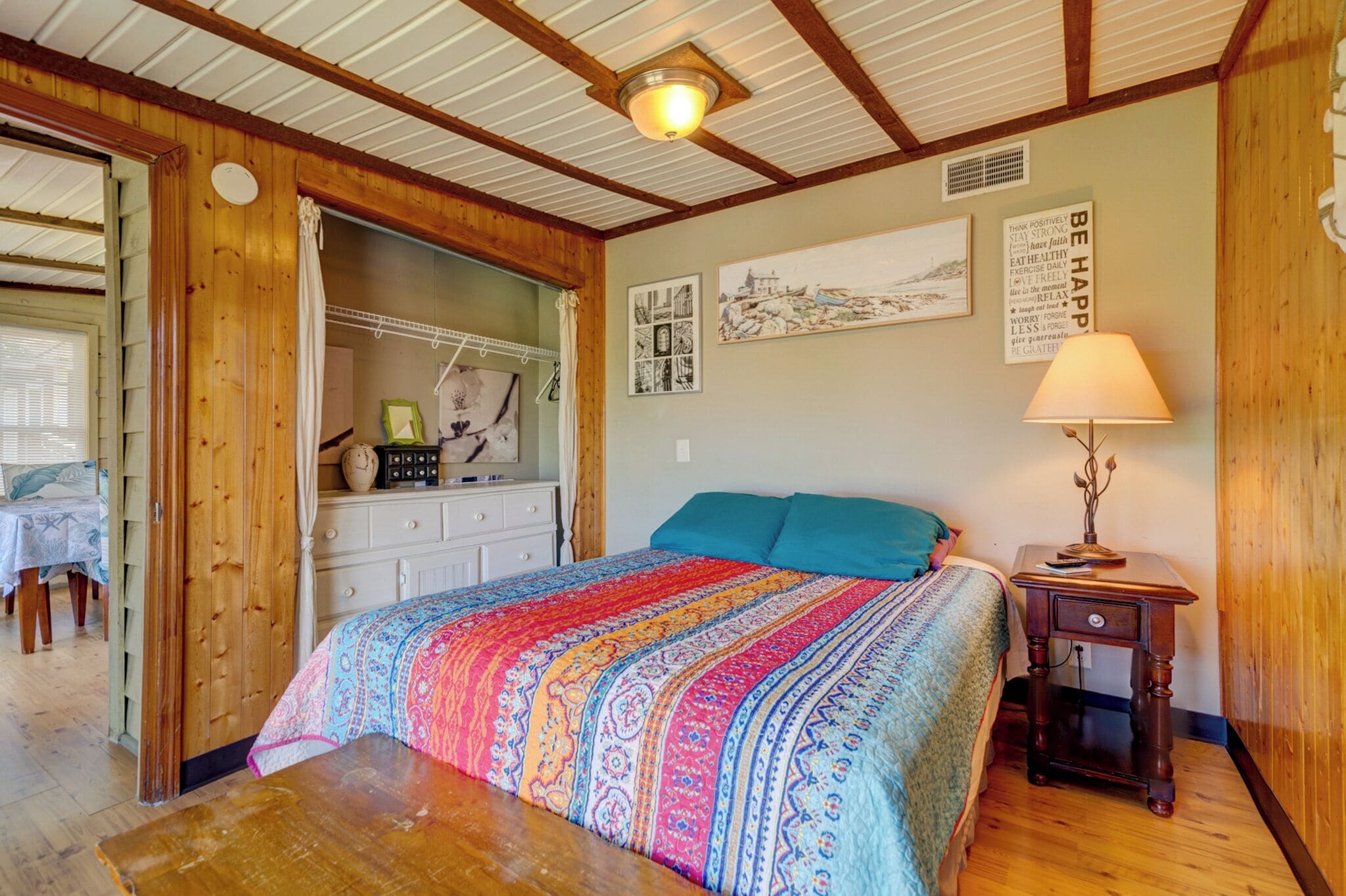 A cozy bedroom with a colorful bedspread, a wooden ceiling, and a small bedside table with a lamp.