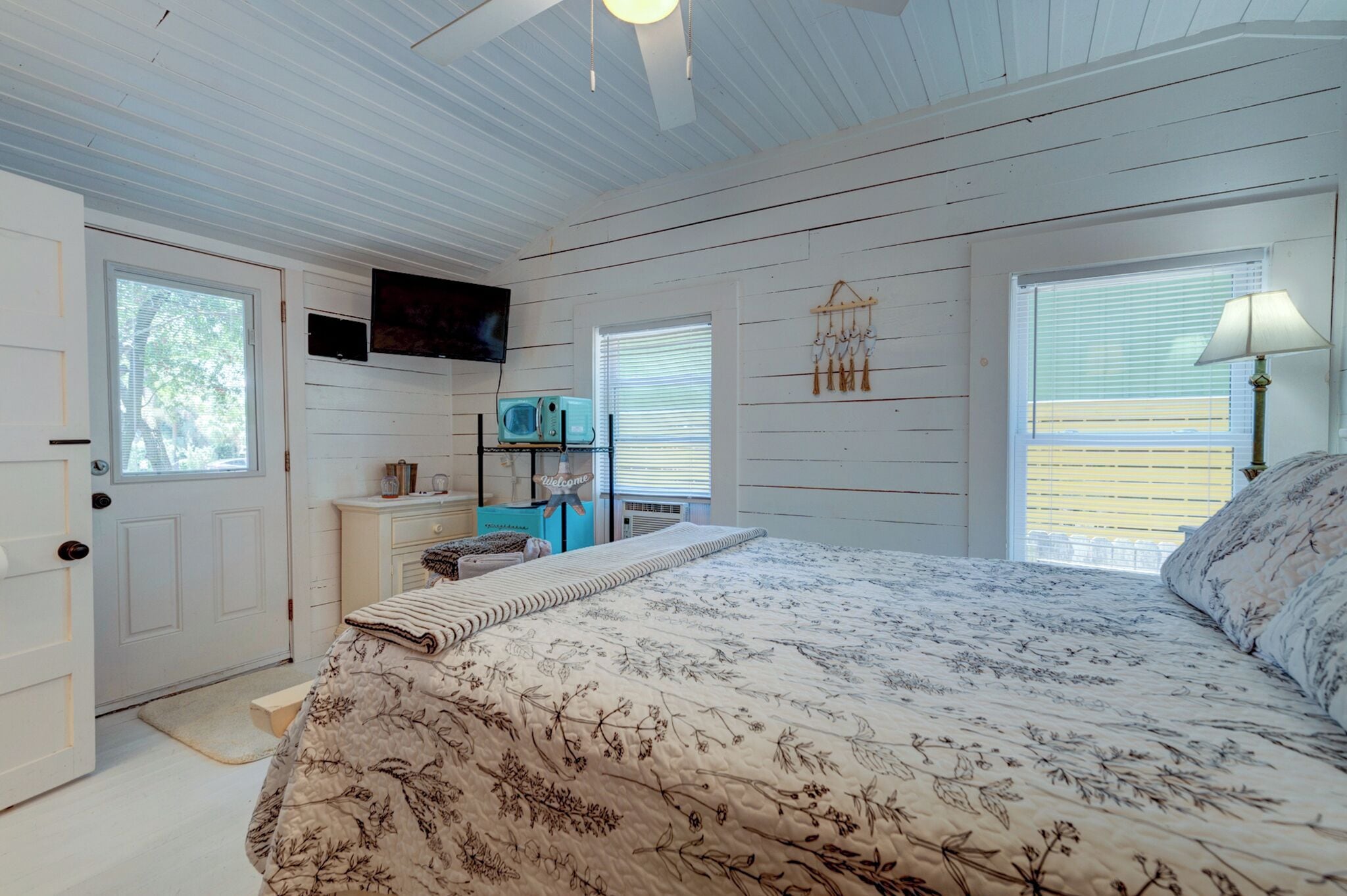 A bedroom with a bed, a TV mounted on the wall, a dresser, and a window with blinds.