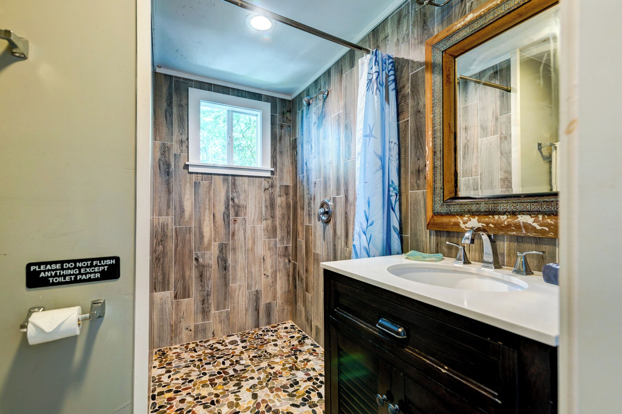 A bathroom with a wooden wall, a shower with a blue curtain, a sink with a mirror, and a sign instructing not to flush toilet paper.