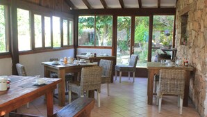 Un comedor rústico con mesas de madera y sillas de mimbre, amplias ventanas con vistas a la vegetación y una pared de piedra.