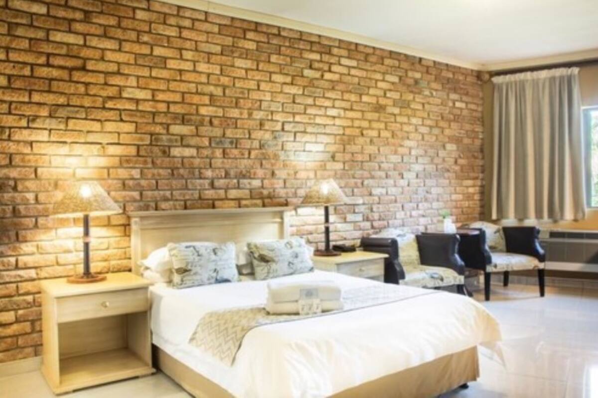 A hotel room with a brick wall, a bed with white linens, two bedside tables with lamps, a chair, and a window with curtains.