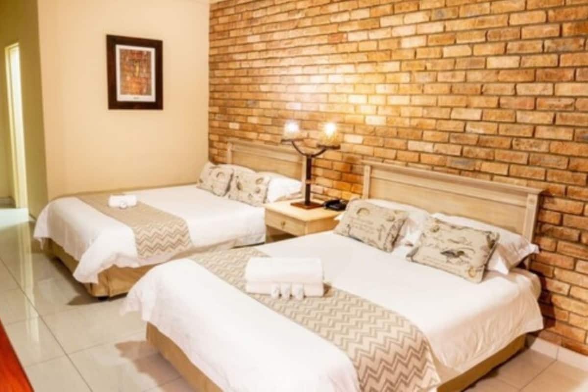 A hotel room with two beds, a brick wall, a framed picture, and a nightstand with a lamp.