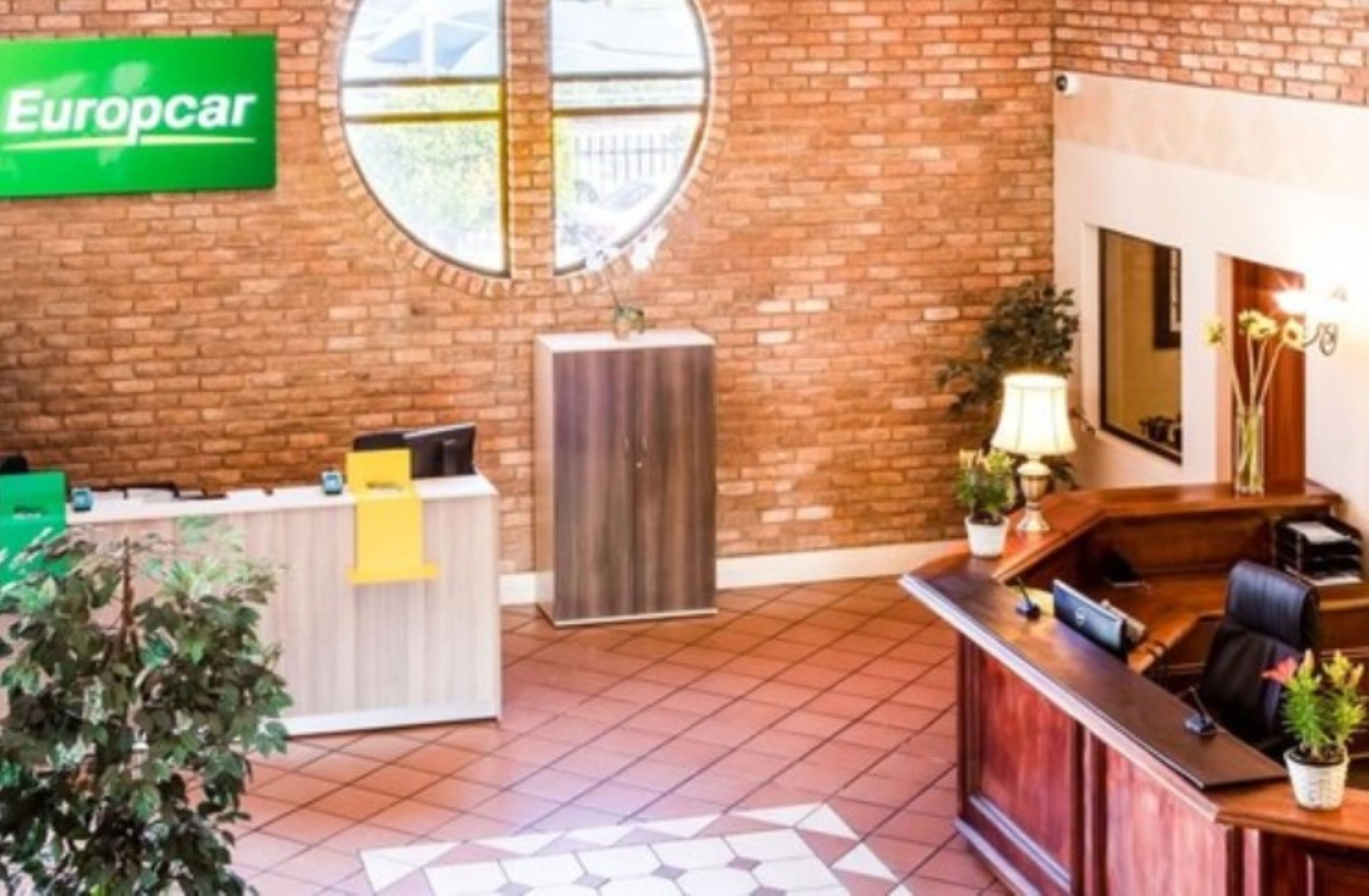 A reception area with a brick wall, a green sign for Europcar, a round window, and a desk with a lamp and plants.