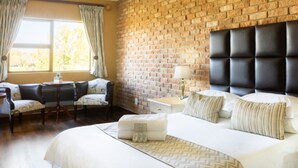 Quarto de hotel com parede de tijolos, uma cama grande com cabeceira, área de estar com sofá e poltronas, e janela com cortinas.