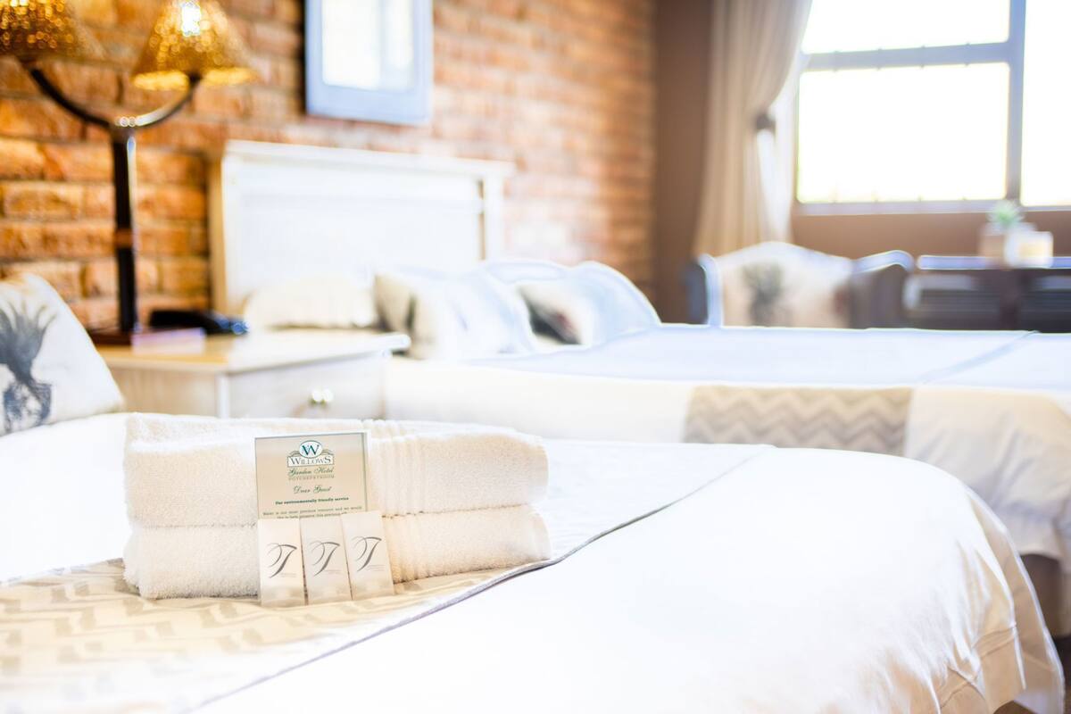 A hotel room with two beds, a brick wall, a lamp, and a window.