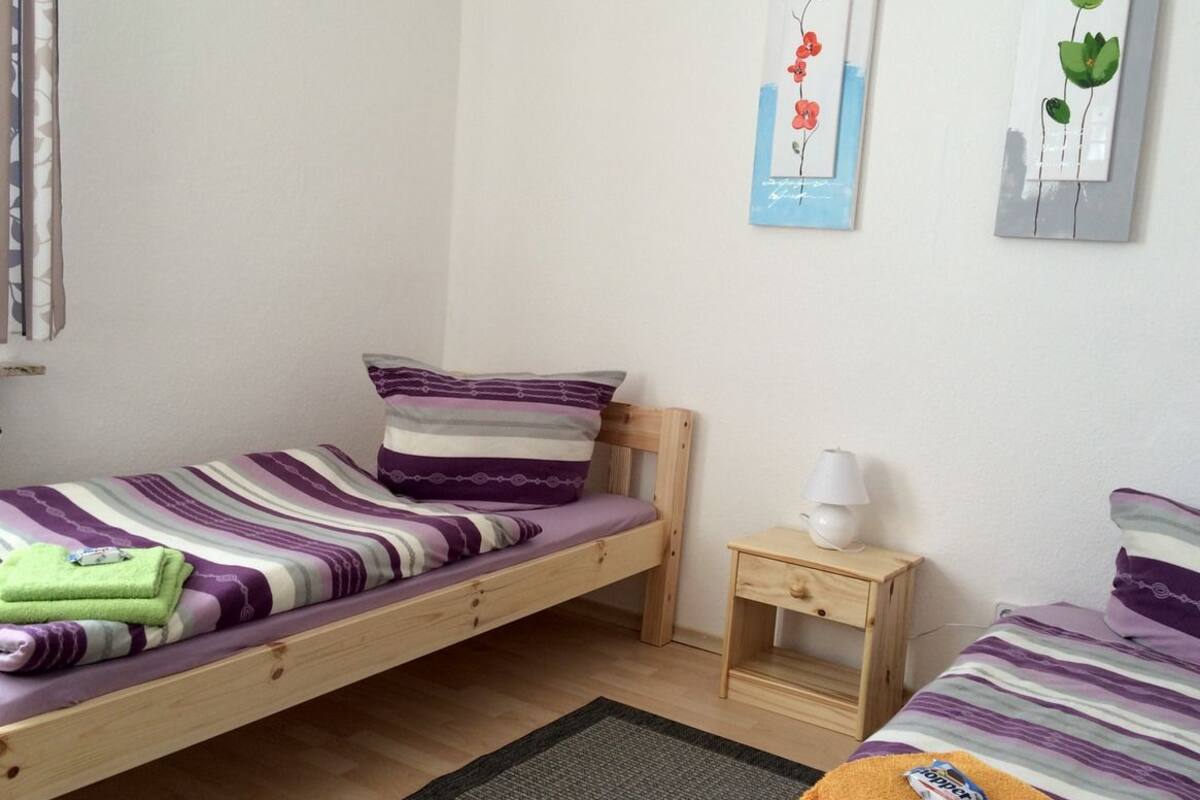 Una habitación pequeña y sencilla con dos camas individuales, una mesita de noche de madera y una lámpara fijada a la pared.