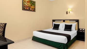 A hotel room with a bed, a desk, a chair, a nightstand, and a wall-mounted lamp.