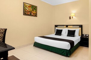 A hotel room with a bed, a desk, a chair, a nightstand, and a wall-mounted lamp.