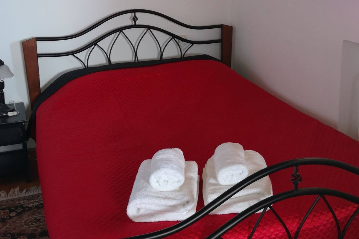 A bed with a red bedspread, a metal headboard, and two rolled towels.