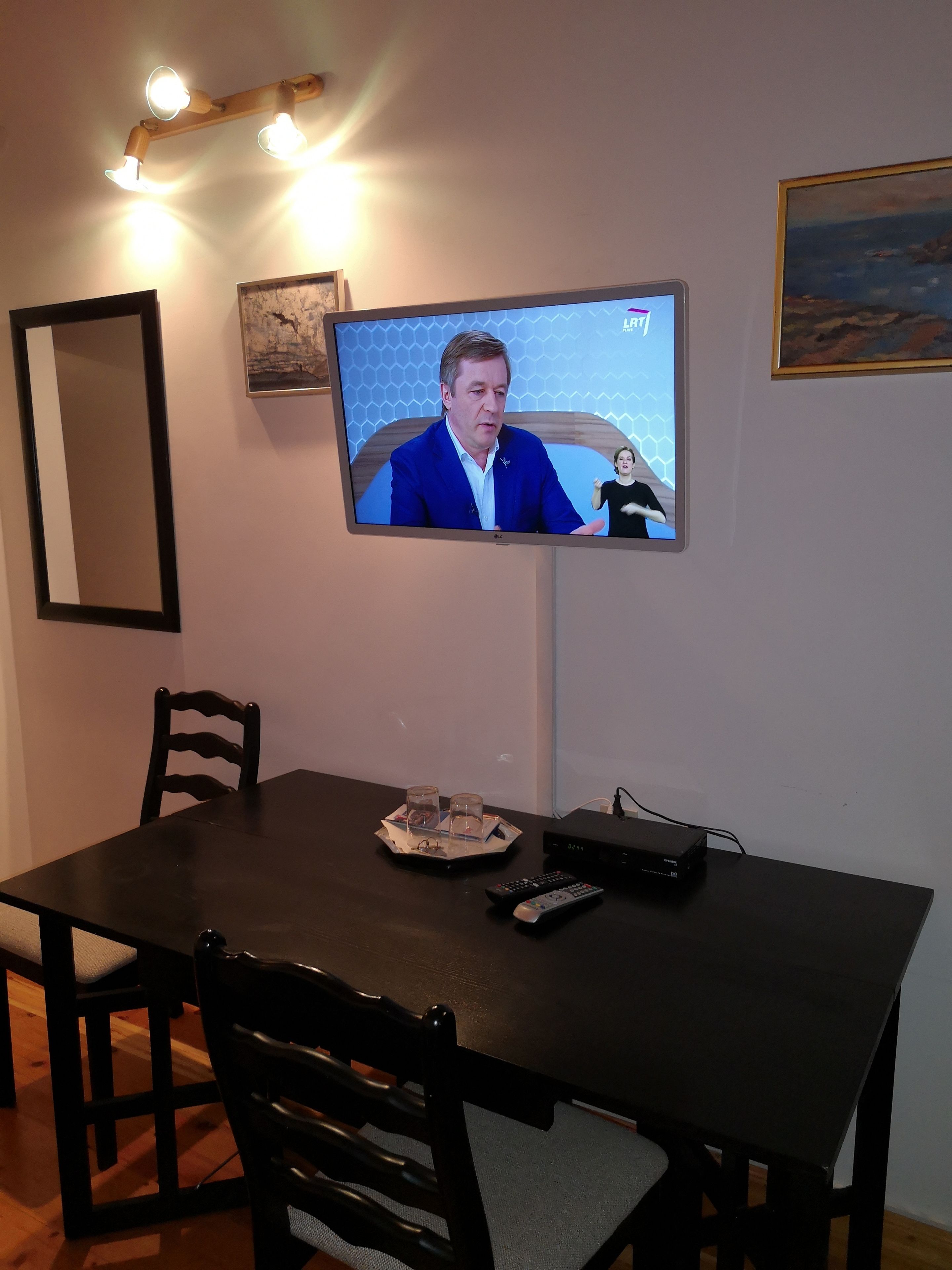 Uma televisão fixada na parede, uma mesa de jantar com cadeiras e um controle remoto sobre a mesa.