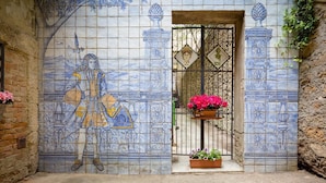 Een betegelde muur met een muurschildering van een figuur met een staf, een poort met een decoratieve sleutel en een bloemenplanter.