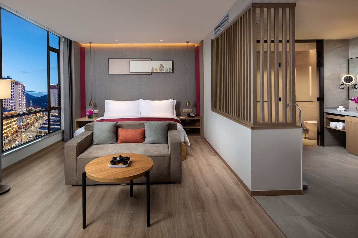 A modern hotel room with a bed, a sofa, a coffee table, and a view of the cityscape.