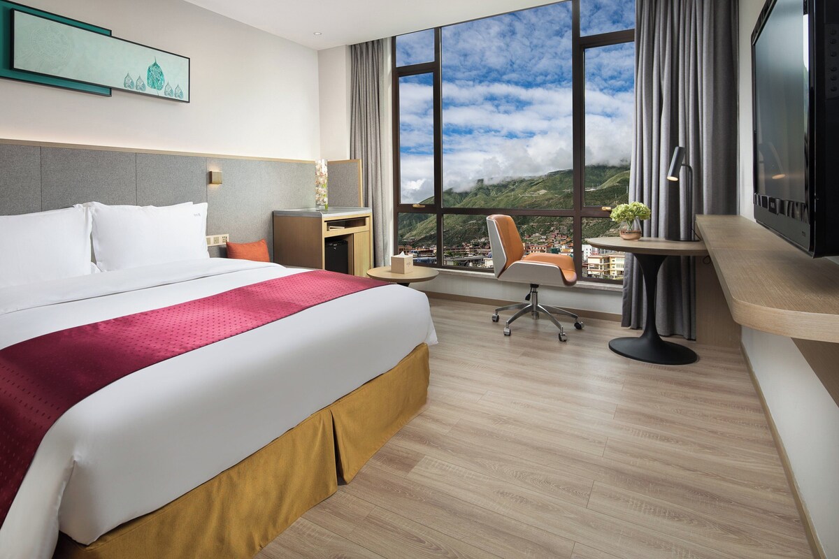 A hotel room with a large bed, a desk, a chair, a TV, and a window with a view of a mountainous landscape.