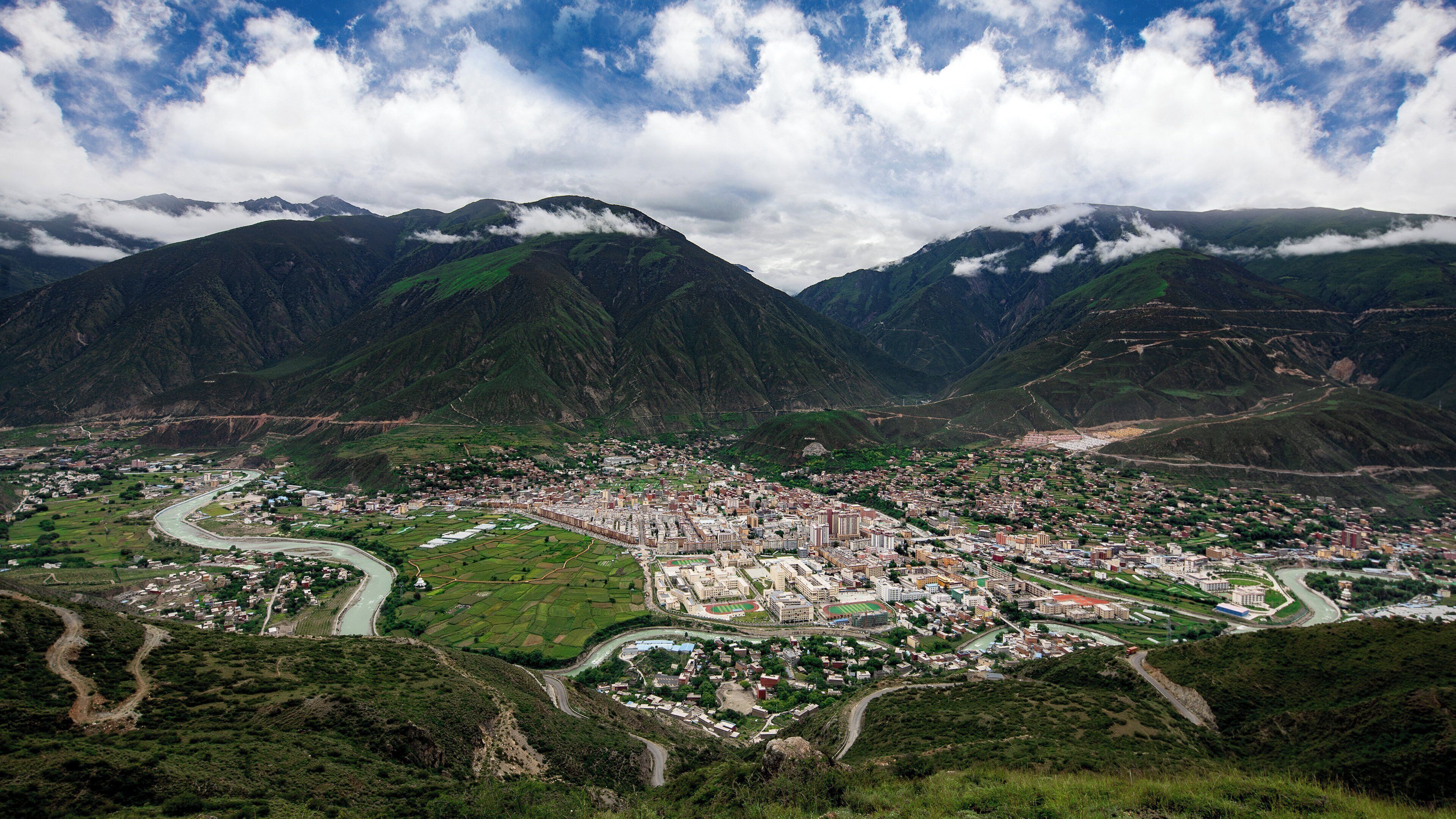 Un paysage montagneux avec une ville nichée dans la vallée, caractérisé par des routes sinueuses, des champs verdoyants et une rivière.