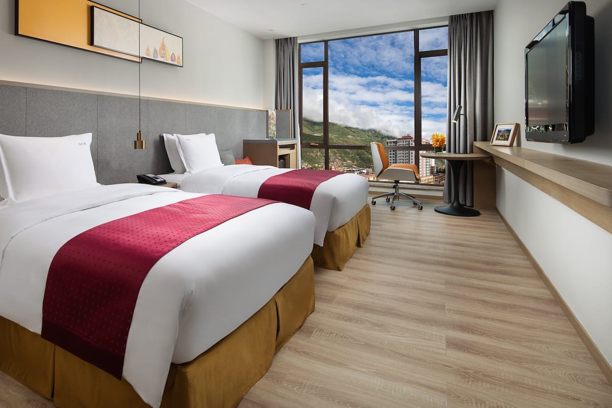 A hotel room with two beds, a desk, a chair, a TV, and a large window with a mountain view.