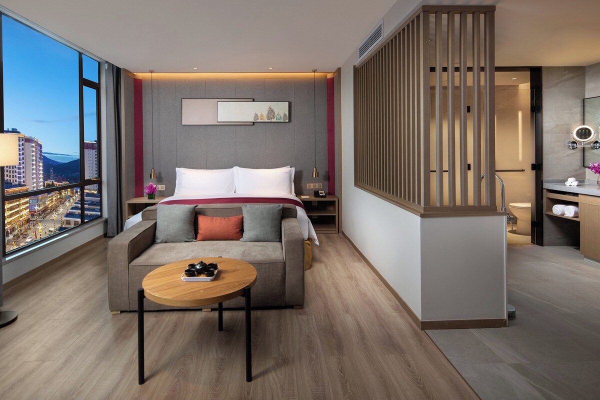 A hotel room with a bed, a sofa, a small table, a nightstand, a bathroom with a mirror and sink, and a large window with a city view.