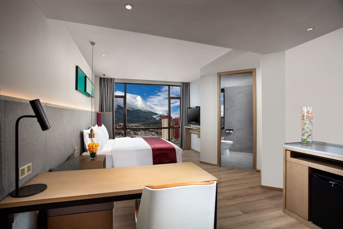 A hotel room with a bed, desk, chair, and a view of a cityscape through large windows.