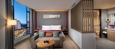 A modern hotel room with a large window, a sofa, a small table, and a bathroom with a mirror and sink.
