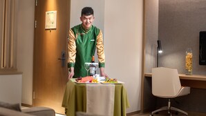 A hotel room with a wooden door, a table set with food, a chair, and a desk with a lamp.