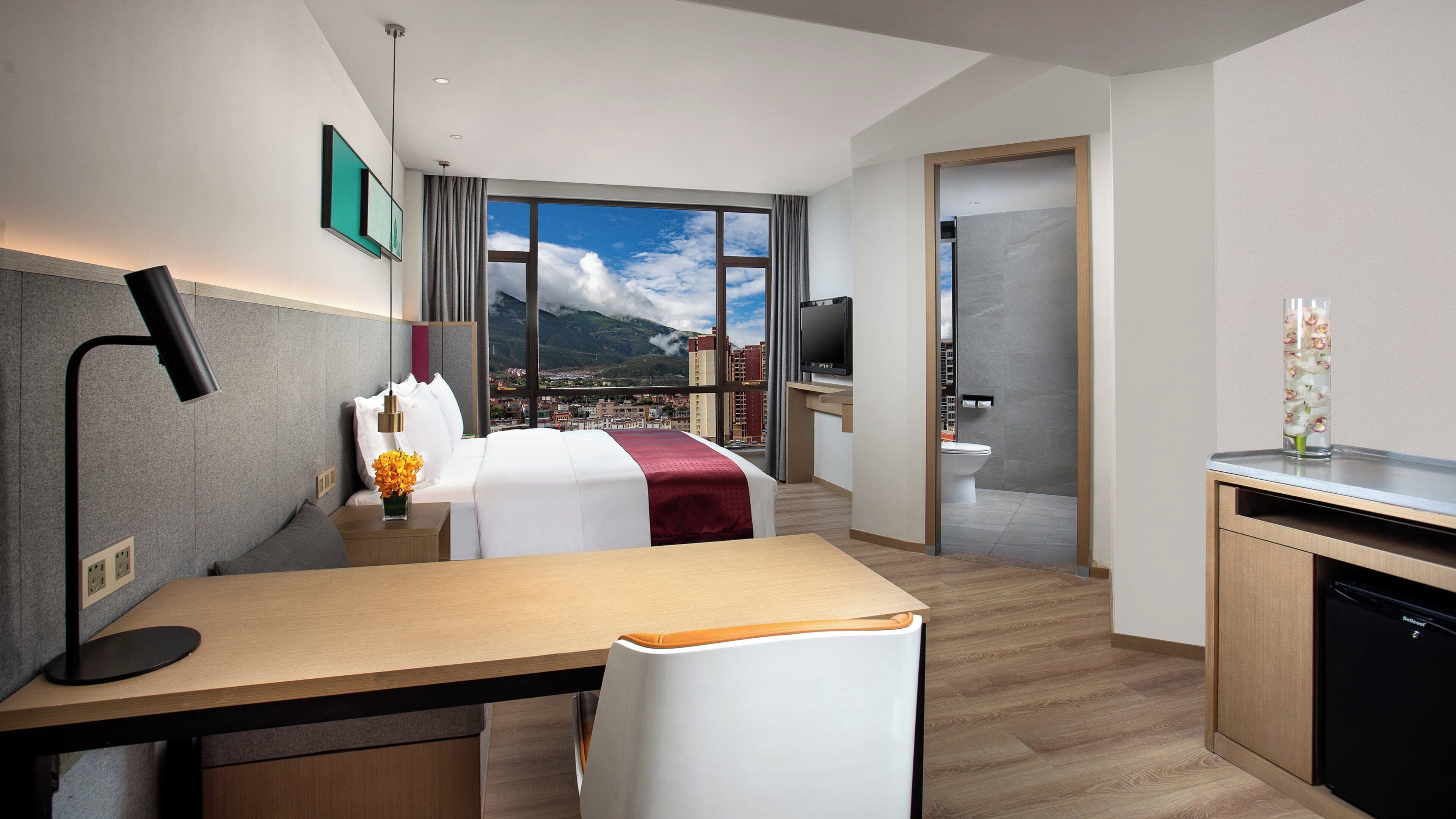 A hotel room with a bed, desk, chair, and a view of a cityscape through large windows.