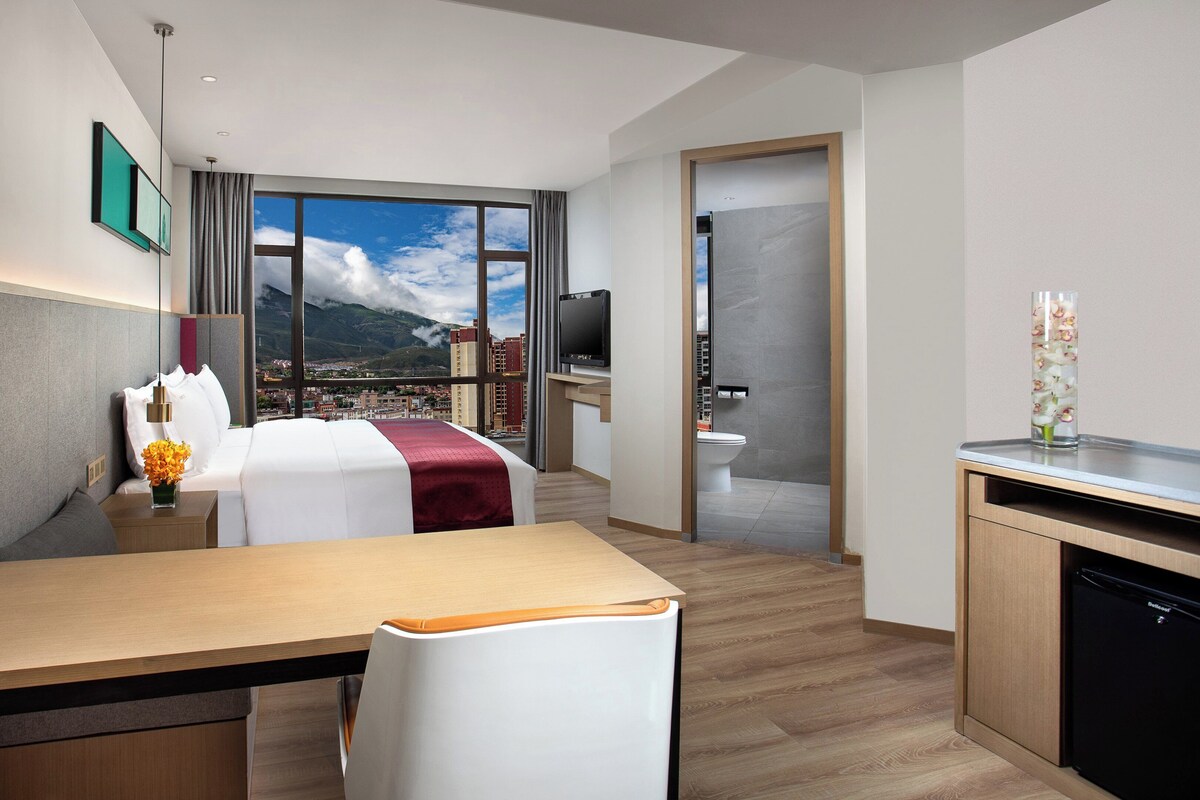 A hotel room with a bed, desk, chair, and a view of a cityscape through large windows.