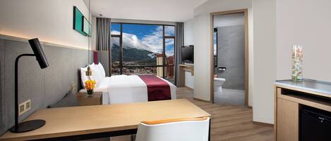 A hotel room with a bed, desk, chair, and a view of a cityscape through large windows.