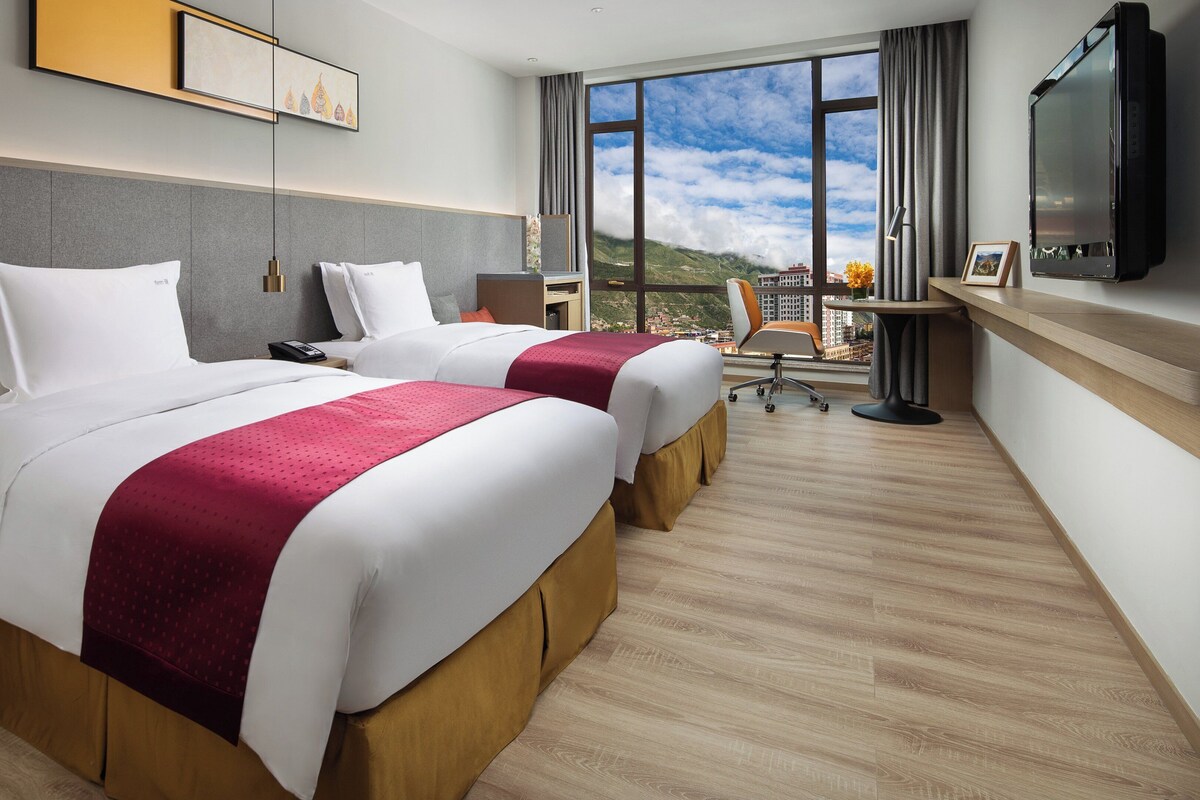 A hotel room with two beds, a desk, a chair, a TV, and a large window with a view of the mountains.