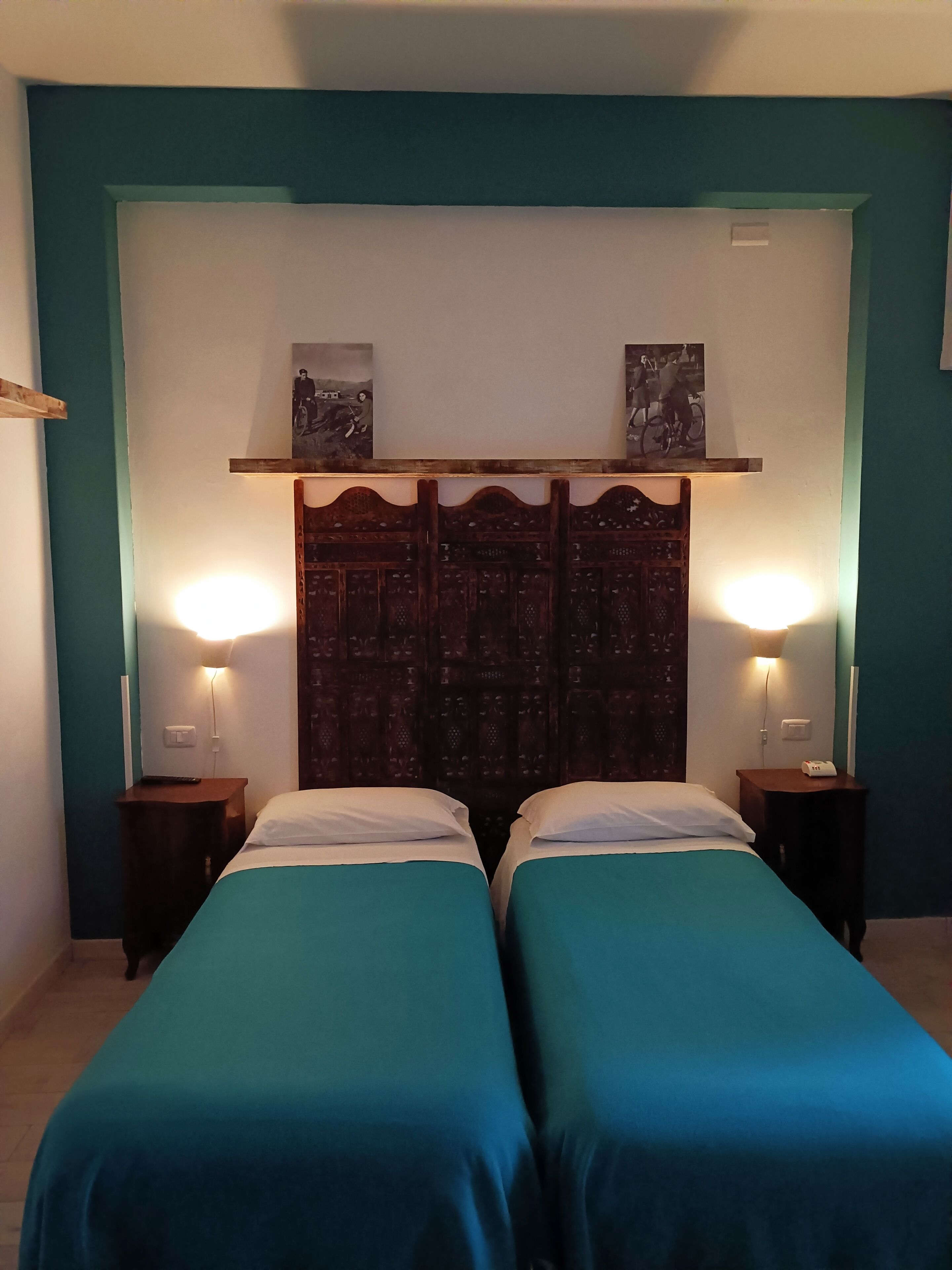 Una habitación con dos camas, ropa de cama color turquesa, un cabecero de madera con detalles artísticos y lámparas de noche.