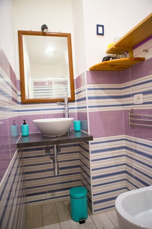 A bathroom with a white sink, mirror, and bathtub. There is a shelf above the sink and a trash can in the corner. - Lanzies Guesthouse (Rome)