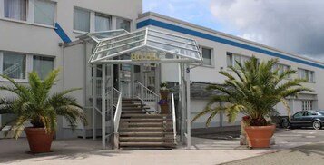 Entrada de hotel con marquesina de vidrio, una escalera y plantas en macetas.