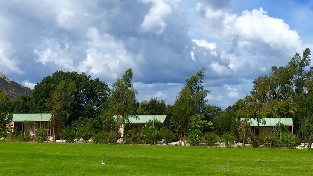 A grassy field with a golf hole marker, surrounded by trees and two buildings with green roofs. - 4/6-person CLIMATIZED WOODEN CHALET, near Algajola beach, countryside & village views (Pigna)