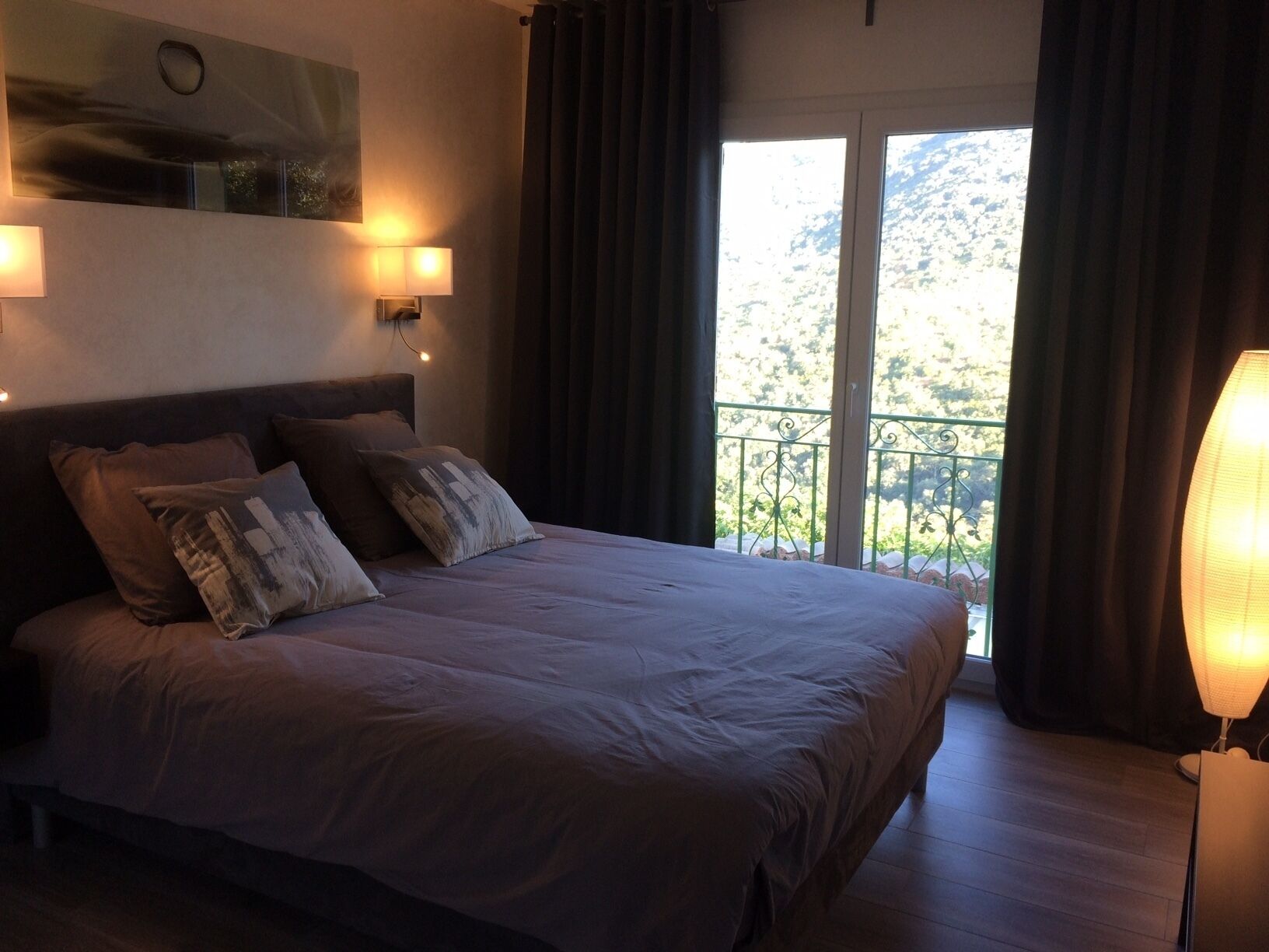 Un dormitorio con una cama grande, dos lámparas de noche, vistas a un balcón y un cuadro en la pared.