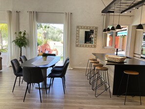 A modern kitchen with a dining area, featuring a large island with stools, a potted plant, and a view of the outdoors.