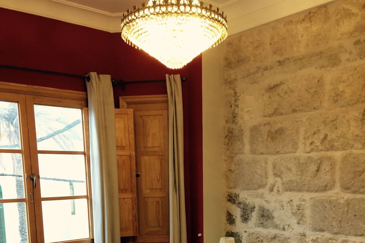 A bedroom with a large bed, a nightstand with a lamp, a stone wall, a wooden door, and a window with curtains.