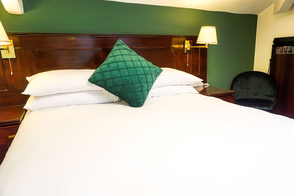 A neatly made bed with white linens and a green quilted pillow, a wooden headboard, and two bedside lamps.