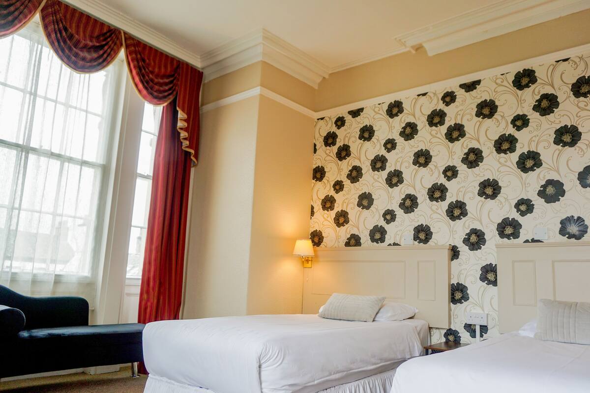 A hotel room with two beds, a sofa, a window with curtains, and a floral wallpaper.