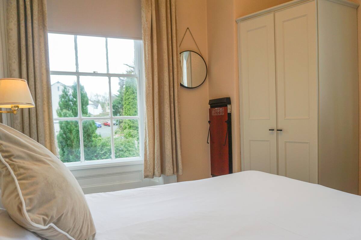 A neatly made bed with white linens and a beige pillow, a window with curtains, a mirror on the wall, and a closed door.
