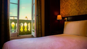A hotel room with a bed, a nightstand with a lamp, a window with curtains, and a view of a building outside. - Manor House Hotel Cockermouth (Cockermouth)